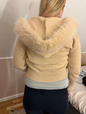 Beige stickad hoodie med fuskpäls, prislapp på - Beige stickad hoodie med dragkedja och stor luva kantad med mjuk fuskpäls. Tröjan har ribbade muddar vid ärmslut och midja, samt en figurnära passform. Perfekt för dig som vill ha en mysig och trendig look.