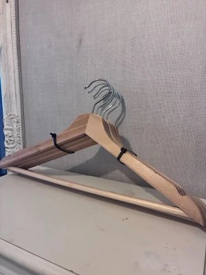 Trägalgar med metallkrok, 5-pack - Säljer ett set med fem klassiska galgar i ljust trä med silverfärgade metallkrokar. Perfekta för att hänga upp kläder i garderoben. Galgarna har en stadig form och en stång för byxor eller kjolar.En av galgarna har tappat stången.