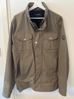 Olivgrön vårjacka från J.Lindeberg - Stilren olivgrön overshirt vårjacka från J.Lindeberg med hög krage, tryckknappar och fyra framfickor. Jackan har JL på ena ärmen och är tillverkad i ett slätt, lätt material som passar perfekt för lager-på-lager. Snygg detalj med metallblixtlås vid halsen.