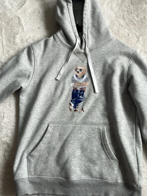  Ralph Lauren hoodie - Säljer en grå hoodie från Polo Ralph Lauren med klassisk huva och vit dragsko. På bröstet finns en broderad björn med cool outfit. Hoodien har en stor magficka och är gjord i mjukt bomullsmaterial. Perfekt för dig som gillar stil och komfort. St M. Men sitter som en s/xs är 174 och den sitter bra