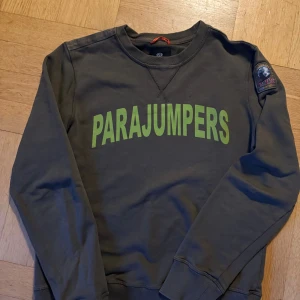 Mossgrön  sweatshirt från Parajumpers - Cool mossgrön sweatshirt från Parajumpers med stort grönt tryck på bröstet och broderad patch på ärmen. Tröjan har rund hals, ribbade muddar och är tillverkad i mjukt material. Perfekt för en avslappnad och trendig look. M ungdoms storlek, ca 170 cl