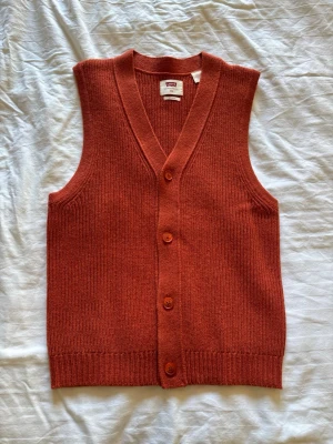 Rostbrun stickad väst med knappar - Snygg rostbrun stickad väst från Levi's med v-ringning och fyra knappar framtill. Klassisk rak passform och ribbade kanter. Perfekt att styla över en skjorta eller t-shirt för en trendig lager-på-lager-look.