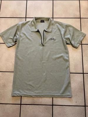 Grå ribbad Oakley t-shirt med half zip, 100% bomull  - Snygg grå t-shirt från Oakley med diskreta ränder och en unik half zip-dragkedja framtill. T-shirten har korta ärmar, liten krage och Oakley-logga broderad på bröstet. Perfekt för dig som gillar sportig och avslappnad stil.