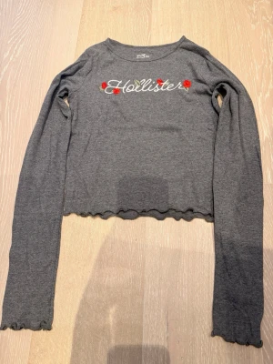 Topp från Hollister  - Grå långärmad topp från Hollister med broderad logga och röda rosor framtill. Croppad modell med vågig kant nertill och vid ärmslut. Tillverkad i mjuk bomullsmix, perfekt för en chill och trendig look.
