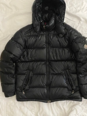 Svart dunjacka från Moncler med huva - Snygg svart dunjacka från Moncler med glansig finish och stor huva. Jackan har två dragkedjeförsedda fickor fram och klassisk Moncler-logga på ärmen. Perfekt för kalla vinterdagar och riktigt varm tack vare dunfyllningen.