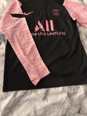PSG Nike träningströja svart/rosa - Snygg PSG träningströja från Nike i svart med rosa ärmar och detaljer. Tröjan har halv dragkedja, Dri-FIT material som andas och klubbmärke på bröstet. Perfekt för fotbollsträning eller gymmet.