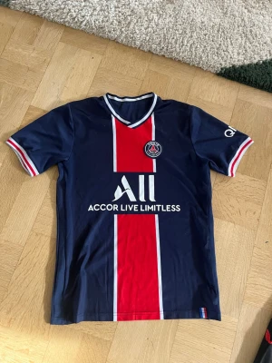 PSG matchtröja blå röd vit - Säljer en snygg PSG matchtröja i mörkblått med röda och vita detaljer. Tröjan har klubbens logga på bröstet, sponsortryck och en röd vertikal rand framtill. Korta ärmar med vita och röda muddar, tillverkad i lätt syntetmaterial.