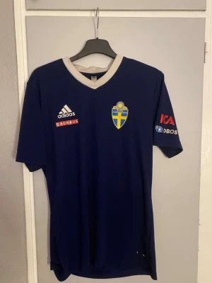 Blå svensk landslagströja Adidas - Säljer en blå svensk landslagströja från Adidas med vit krage och korta ärmar. Tröjan har Svenska Fotbollförbundets emblem på bröstet samt sponsorloggor från Bauhaus, ICA och OBOS. Perfekt för fotbollsfans som vill visa sitt stöd.