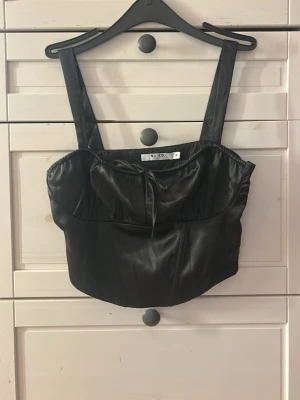 Svart topp från NA-KD - Snygg svart korsett-topp från NA-KD med breda axelband och knyt framtill. Toppens material har en lätt glansig finish som ger en cool look. 🥰