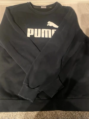 Svart sweatshirt från Puma - Klassisk svart sweatshirt från Puma med stor vit logga och text på bröstet. Tröjan har rund halsringning, ribbade muddar vid ärmslut och nederkant samt en avslappnad passform. Perfekt för en sportig och casual stil. Pris kan diskuteras. 