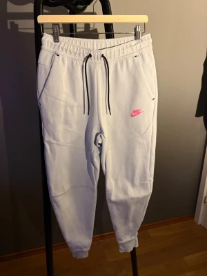 Vita mjukisbyxor från Nike - Snygga vita mjukisbyxor från Nike med svart dragsko i midjan och rosa Nike-logga på vänster ben. Byxorna har två sidofickor och muddar vid bensluten. Perfekta för en avslappnad och sportig stil.