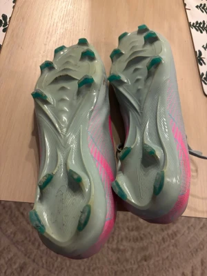 Nike Mercurial grå/rosa fotbollsskor pro model - Snygga Nike Mercurial fotbollsskor i grått med rosa detaljer och logga. Skorna har en smal, aerodynamisk form och är tillverkade i syntetmaterial med fasta dobbar för spel på gräs. Perfekta för dig som vill ha snabbhet och stil på planen.