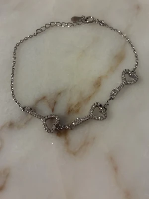 Silverarmband med hjärtan och stenar - Ett tunt silverfärgat armband med tre hjärtformade länkar dekorerade med små glittrande stenar. Perfekt för dig som gillar söta detaljer och stilren design. Justerbar längd med karbinlås.