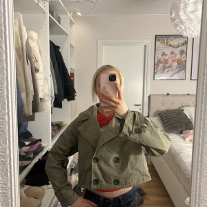 Kort trenchcoat från Zara i khaki - Snygg kort trenchcoat från Zara i khakigrönt bomullstyg. Jackan har dubbelknäppning framtill, bred krage och en croppad passform som ger en trendig look. Perfekt att slänga över en hoodie eller t-shirt för en cool streetstyle.