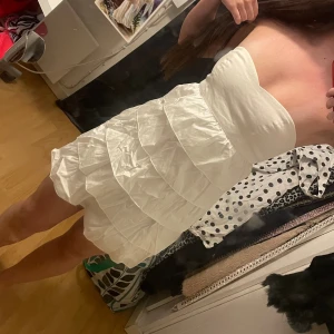 Vit axelbandslös kortklänning - Axelbandslös vit kortklänning med volanger och dragkedja i ryggen. Klänningen har en snygg, figurnära passform och är perfekt för dig som vill ha en stilren och trendig look. Materialet känns lätt och passar bra till varmare dagar.