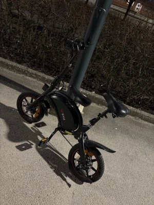 Svart elcykel från DYU - Säljer en kompakt och smidig svart elcykel från DYU med rundad ram och små hjul. Cykeln har en bekväm sadel, eldrift och laddningsuttag på sidan. Perfekt för dig som vill ta dig fram snabbt i stan. Modern design med röda detaljer på däcken. Den funkar exakt som den ska men bromsarna kan vara lite lite sega och har tyvärr inte laddare till den, och om nån ska köpa får de hämtäs 