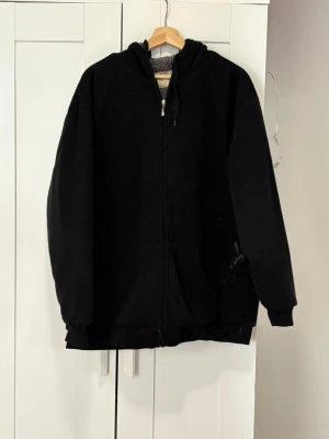 Svart hoodie med dragkedja och huva - En klassisk svart hoodie med dragkedja framtill och stor huva. Hoodien har två fickor på sidorna och ribbade muddar vid ärmslut och nederkant. Perfekt för en avslappnad och stilren look. Materialet ser ut att vara mjukt och bekvämt, troligen bomullsblandning. Pris går att diskutera 