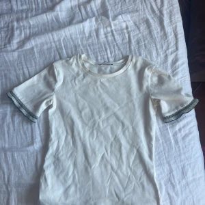 Vit t-shirt med ribbade ärmslut - Vit t-shirt med korta ärmar och ribbade muddar i grått på ärmsluten. Enkel och stilren design som passar perfekt till jeans eller shorts. Materialet känns mjukt och bekvämt, troligen bomull. Snygg detalj med kontrastfärg på ärmarna.