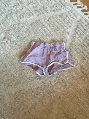 Ljuslila shorts med vita kanter - Säljer ett par ljuslila shorts med vita kantband och elastisk midja. De har en sportig design och är perfekta för varma dagar. Materialet känns mjukt och lätt, vilket gör dem bekväma att bära. Passformen är loose och de har en klassisk look.