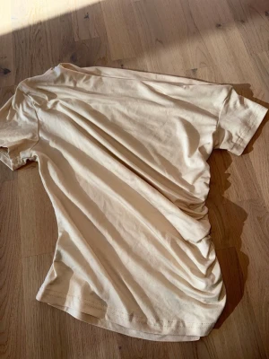Beige topp med rynkade detaljer - Säljer en beige topp med korta ärmar,sne skuren och snygga rynkade detaljer på sidan. T-shirten har en enkel och clean look, perfekt för att matcha med jeans eller kjol. Materialet är mjukt och känns skönt mot huden.