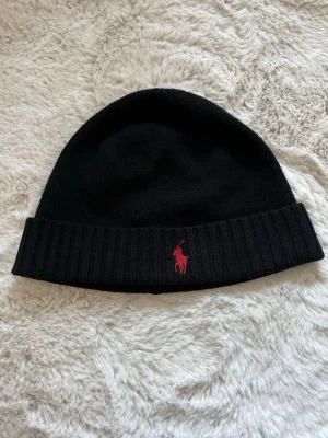 Mössa Polo Ralph Lauren - Snygg svart mössa från Polo Ralph Lauren i mjuk merinoull. Klassisk ribbad kant och ikoniska röda loggan broderad framtill