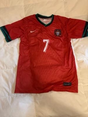 Portugal fotbollströja Nike röd - Röd Portugal fotbollströja från Nike med nummer 7 och namn på ryggen. Tröjan har gröna detaljer vid krage och ärmslut samt broderad Portugal-logga på bröstet. Tillverkad i lätt och ventilerande material, perfekt för träning eller match.
