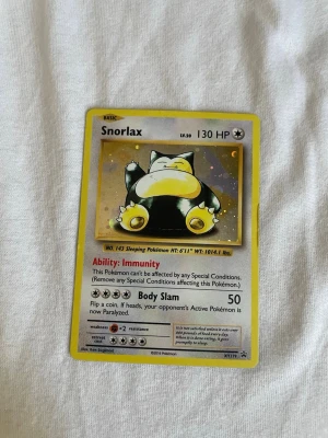 Snorlax Pokémonkort XY178 - Snorlax Pokémonkort från XY Black Star Promos, nummer XY178. Kortet har gul ram, färgglad illustration av Snorlax och detaljer om förmåga och attack. Baksidan har klassisk blå Pokémon-design med pokéboll. Perfekt för samlare eller Pokémonfans.
