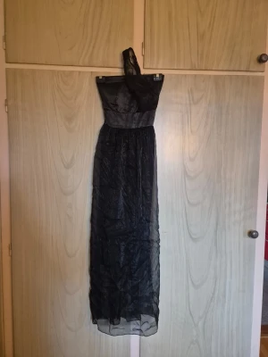 Svart långklänning oneshoulder - Elegant svart långklänning i oneshoulder-modell med glansigt tyg upptill och ett skirt, lätt transparent tyg nedtill. Klänningen har markerad midja och en axel, vilket ger en stilren och modern look. Perfekt för speciella tillfällen.     Alla kläder rollas innan dom skickas hem då det finns katt i hemmet🐈 Jag stryker och tvättar INTE innan jag skickar det får du stå för själv så det blir som du vill ha det🤗