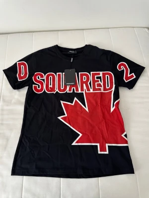 Svart Dsquared2 t-shirt med lönnlöv - Cool svart t-shirt från Dsquared2 med stor röd lönnlövsgrafik och vit-röd text på bröstet. Klassisk rund hals och korta ärmar. Perfekt statement-plagg för dig som gillar streetwear och snygga tryck.