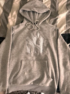 Grå hoodie med NY-logga - Snygg grå hoodie med stor NY-logga i vitt och grått tyg på bröstet. Klassisk känguruficka framtill och justerbar huva med dragsko. Perfekt för en avslappnad streetwear-look.