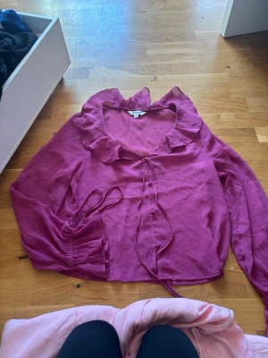 Lila blus med volang från Kappahl - Supersöt lila blus från Kappahl med volangkrage och knytband framtill. Blusen har långärmade ärmar med resår vid handlederna och är gjord i ett tunt, lätt material med struktur. Perfekt för att lyfta vilken outfit som helst!