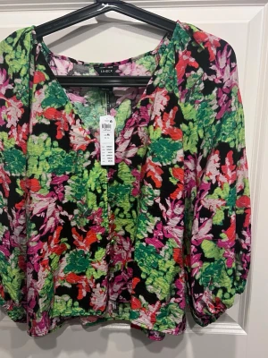 Färgglad blus med knappar från LinDEX - Blus från LinDEX med färgstarkt blommönster i grönt, rosa, rött och svart. V-ringad med dekorativa knappar framtill och långärmad modell med lätt puff vid ärmslut. Tillverkad i ett tunt, strukturerat tyg som ger en luftig känsla.