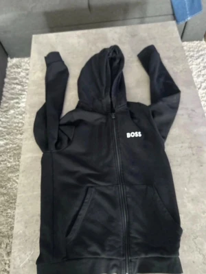 Svart hoodie från BOSS med dragkedja - Svart hoodie från BOSS med vit logotyp på bröstet. Hoodien har dragkedja framtill, två stora fickor och en klassisk huva. Materialet ser ut att vara mjukt och bekvämt, perfekt för en avslappnad och cool look.