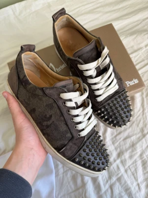  Camo Christian Louboutin skor - Säljer ett par svarta sneakers från Christian Louboutin med vita snören och ikoniska nitar på tån. Alla nitar är kvar. Nypris:11000 mitt pris:3899 Skorna har en låg silhuett och är tillverkade i mocka och läder. Snygg kontrast mellan svart ovandel och vit sula. Perfekta för dig som gillar edgy detaljer. Storlek 41 passar även 42 lådan tillhör ett par andra skor men kan skicka med vid önskan