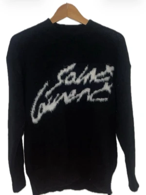 Svart stickad tröja Saint Laurent - Cool svart stickad tröja från Saint Laurent med vit, suddig logotyp framtill. Tröjan har rund halsringning och långa ärmar. Perfekt för dig som gillar streetstyle och vill sticka ut med en statement piece.