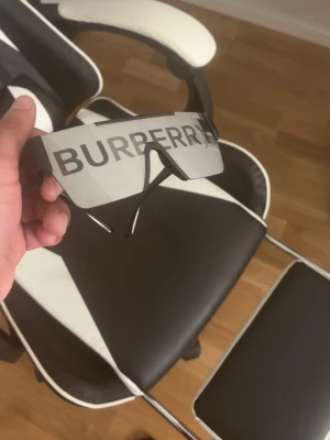 Burberry solglasögon - Säljer ett par futuristiska solglasögon från Burberry med stor, rektangulär sköldformad lins i silver och svart. Glasögonen har en tydlig Burberry-logga över hela linsen och tunna svarta skalmar. Perfekt statement piece för dig som vill sticka ut.