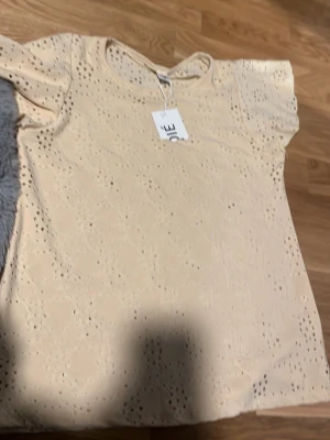 Beige blus med broderade hål från Soié - Söt beige blus storlek M. Toppen har korta ärmar och är dekorerad med broderade hål som ger ett luftigt och trendigt intryck. Perfekt för dig som gillar romantiska detaljer och ljusa färger.