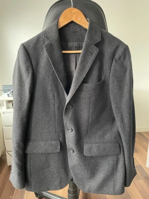 Mörkgrå ullkavaj med klassisk passform - Stilren mörkgrå kavaj i ull med klassisk passform och tre knappar framtill. Kavajen har två fickor med lock och en bröstficka. Perfekt för dig som vill ha en tidlös och snygg look till vardags eller mer uppklätt.