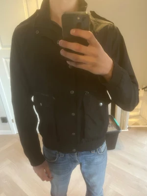 Svart vindjacka med hög krage - Snygg svart vindjacka från Samoe med hög krage och dragkedja samt knappar framtill. Jackan har två fickor med dragkedja och tryckknapp, elastisk nederkant och diskret logotyp på ryggen. Materialet är lätt och glansigt, perfekt för blåsiga dagar. Öppen för byten också!