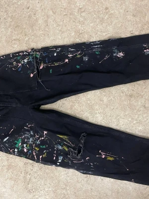 Svarta jeans med färgstänk och slitningar - Unika svarta jeans med coola färgstänk i grönt, rosa, vitt och gult. Jeansen har en avslappnad passform och slitningar på ena benet för en edgy look. Perfekt för dig som vill sticka ut och gillar streetstyle. Materialet är denim.