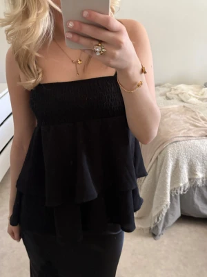 Svart axelbandslös volangtop - Säljer en svart axelbandslös topp med volanger och smock upptill. Toppen har en luftig och flowy passform som ger en snygg siluett. Perfekt att styla med smycken för en trendig look.