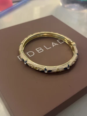 Guldigt armband  - Snyggt armband i guldfärgad metall med vit emalj och små blå detaljer. Armbandet har en oval form och öppnas med ett gångjärn. Perfekt accessoar för att lyfta din outfit med en elegant touch.