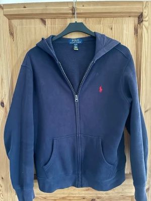 Marinblå hoodie från Polo Ralph Lauren - Säljer en marinblå hoodie från Polo Ralph Lauren med dragkedja, huva och två fickor framtill. Klassisk röd logga broderad på bröstet. Tillverkad i mjukt bomullsmaterial som är skönt mot huden. Perfekt för en chill och stilren look.