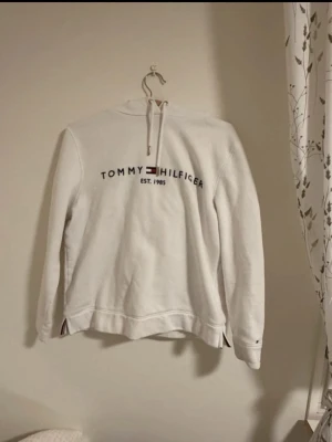 Vit hoodie från Tommy Hilfiger - Vit hoodie från Tommy Hilfiger med broderad logga och text 'EST. 1985' på bröstet. Klassisk huva med dragsko och långa ärmar. Materialet är mjukt och mysigt.