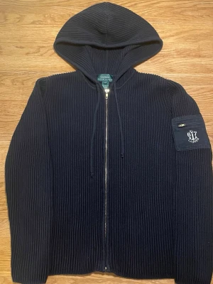 Marinblå stickad hoodie från Lauren Ralph Lauren - Snygg marinblå stickad hoodie från Lauren Ralph Lauren i 100% bomull. Tröjan har dragkedja framtill, huva och en ficka med detalj på ärmen. Perfekt för en chill och stilren look.