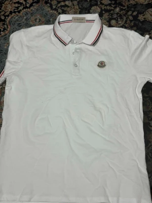 Vit Moncler pikétröja med röd detalj - Snygg vit pikétröja från Moncler med klassisk krage och knäppning framtill. Tröjan har röda och marinblå ränder på kragen och ärmsluten samt Moncler-logga broderad på bröstet. Tillverkad i mjuk bomull som känns skön mot huden.