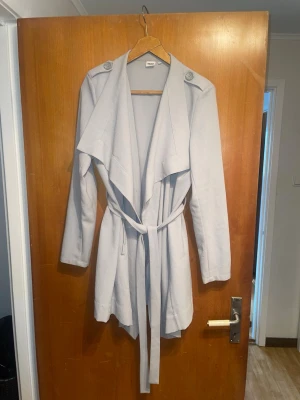 Ljusgrå trenchcoat från Object - Stilren ljusgrå trenchcoat från Object med knytskärp i midjan och breda slag. Jackan har axelklaffar med knappar och ett mjukt, tunt tyg som ger ett snyggt fall. Perfekt för dig som gillar minimalistisk och modern stil.