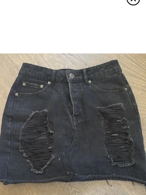 Svart jeanskjol med slitningar - Cool svart jeanskjol med råa slitningar framtill och klassisk femficksdesign. Kjolen är kort och har en rak passform med knapp och dragkedja i midjan. Perfekt för dig som gillar en edgy och trendig stil.