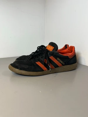 Adidas Spezial svarta och orange sneakers - Snygga Adidas Spezial sneakers i svart mocka med orange detaljer och klassiska tre ränder på sidan. Skorna har lågt skaft, bruna gummisulor och orange logga på plösen. Perfekta för dig som gillar retrostil och streetwear.