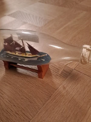 Skepp i flaska med träställ - Dekorativt skepp i flaska med brunröda segel och detaljer i trä och blått. Flaskan är genomskinlig och står på ett litet träställ. Perfekt som prydnad eller samlarobjekt för dig som gillar marina teman.